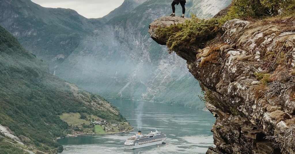 Person enjoys breathtaking view from a cliff above Geiranger Fjord, Norway, with clear skies and cruise ship below.